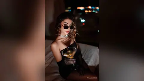 Glamorous woman in a black dress and sunglasses holding a cocktail in a moody nighttime setting.