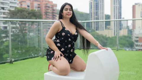 Curvy woman in a black floral romper sitting on a white chair on a rooftop garden with city buildings background.