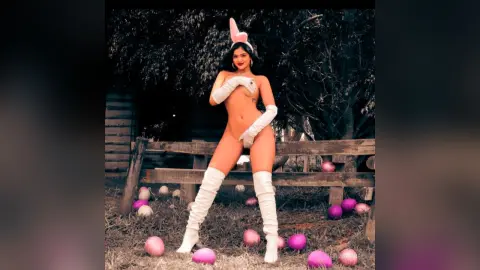 Woman in white bunny ears and gloves posing in a whimsical garden setting with pink pastel eggs.