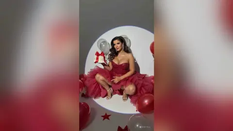 Glamorous woman in a red tulle gown sitting with balloons and a gift box in a festive celebratory setting.