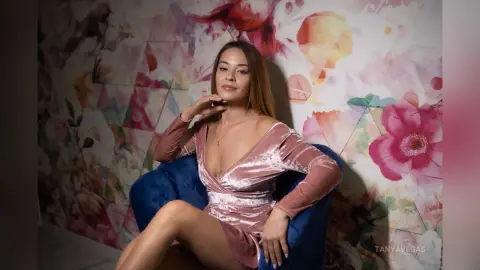 Elegant woman in a pink velvet dress posing against a floral wallpaper backdrop in a soft, romantic mood.