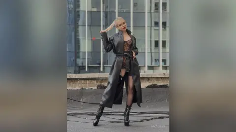 Stylish woman in a long black leather trench coat and leopard print top posing confidently in a city setting.
