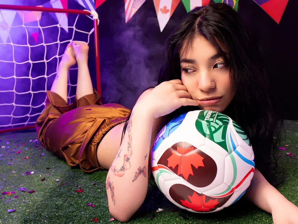Young woman in satin shorts lying on turf with a soccer ball near a goal net in a moody, purple-lit setting.