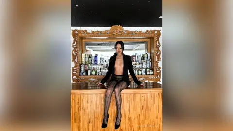 Elegant woman in a black blazer and sheer tights posing at a wooden bar in a moody lounge setting.