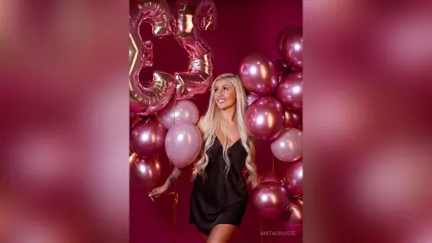 Anita smiles while wearing a black silk slip dress, surrounded by pink and gold balloons against a pink background.