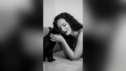 Smiling woman with curly hair cuddling a black cat in a cozy, moody black and white indoor setting.