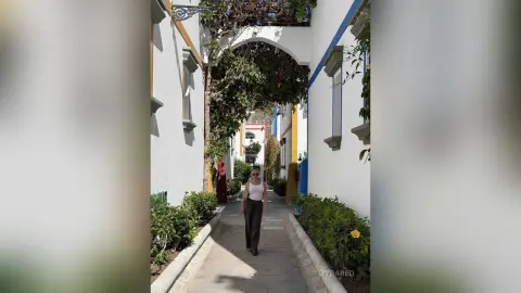 Stylish woman in a white tank top and brown trousers walking through a sunny, narrow Mediterranean alleyway.