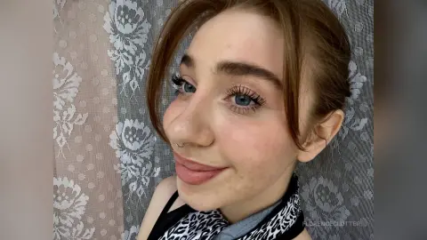 Smiling young woman with long eyelashes and nose ring wearing a leopard print top in front of lace curtains.