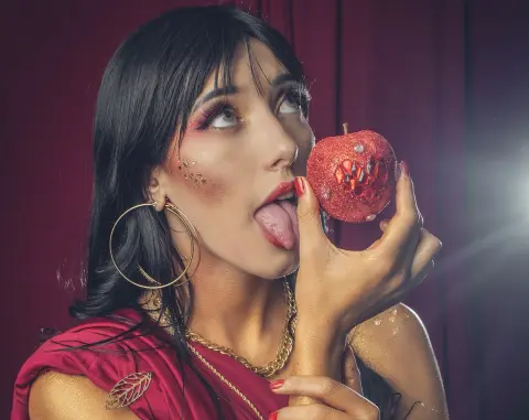 Glamorous woman in red outfit biting a glittery red apple against a dark moody background.