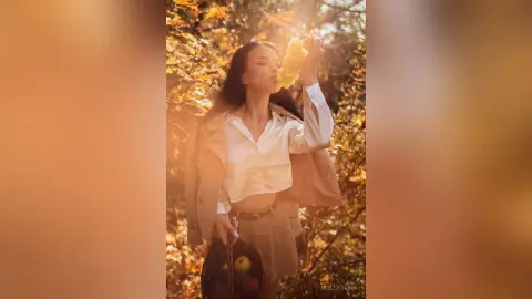 Young woman in a white shirt and beige blazer smelling autumn leaves in a golden sunlit forest setting.