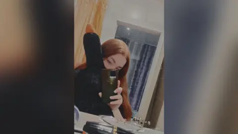 Redheaded woman in a black long-sleeve top taking a moody mirror selfie in a modern bathroom.