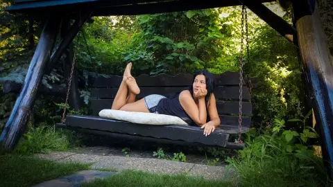 MissyJolie reclines on a dark wooden swing in a lush green garden, wearing a black tank top and grey shorts.