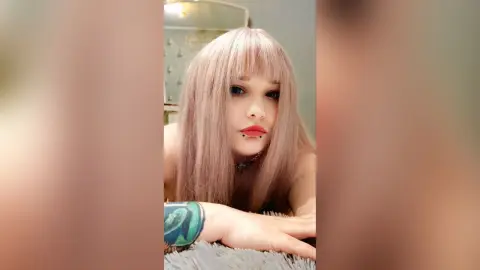 EstherTrix with long pink hair and a black choker poses closely for a selfie while lying down.