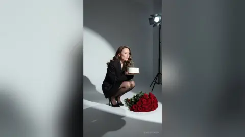 EmilyGiffin squats in a black blazer and sheer black tights, holding a small white cake with a single lit candle next to a pile of red roses.