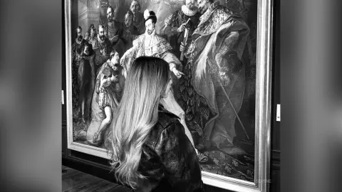 Woman with long hair in a leather jacket viewing a large classical historical portrait in a museum gallery.