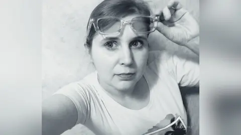 Close-up portrait of a woman in a white t-shirt wearing clear glasses with a casual, neutral expression.
