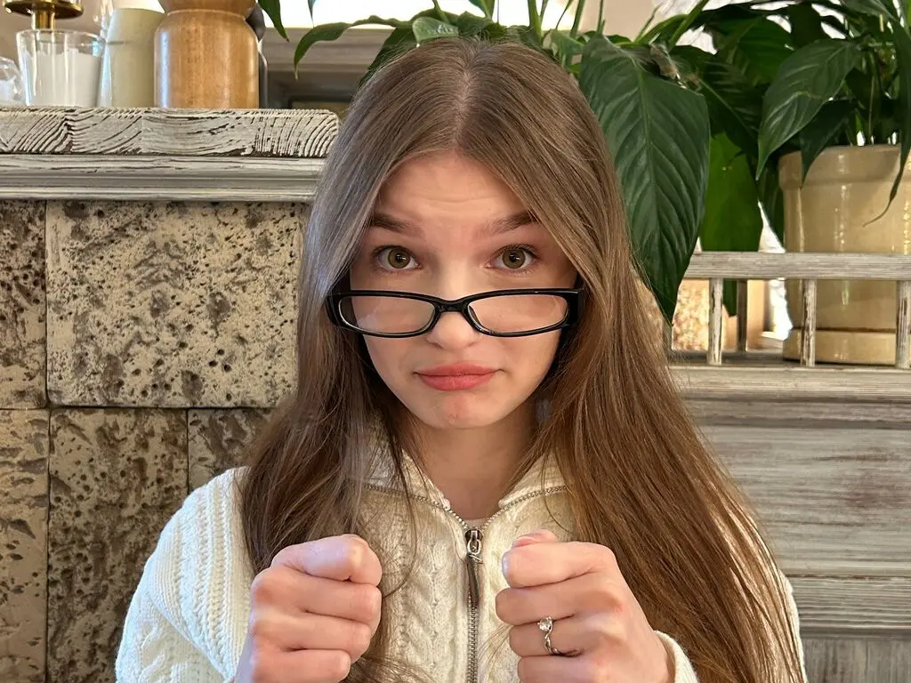 Young woman with long brown hair and glasses wearing a white knit sweater in a bright indoor setting.