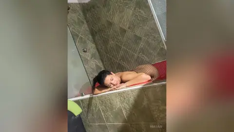 Young woman in a beige bodysuit relaxing on a red mat in a modern green tiled bathroom.