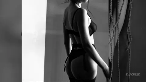 Moody black and white portrait of a woman in black lingerie posing in a minimalist studio setting.