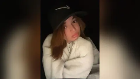 Young woman in a cozy white fleece hoodie and black Nike baseball cap posing in a moody, dark setting.