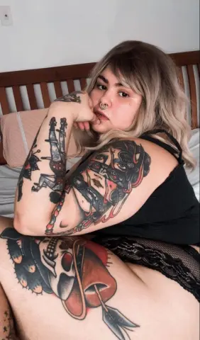 Woman with blonde hair and extensive arm tattoos wearing a black tank top in a cozy bedroom setting.