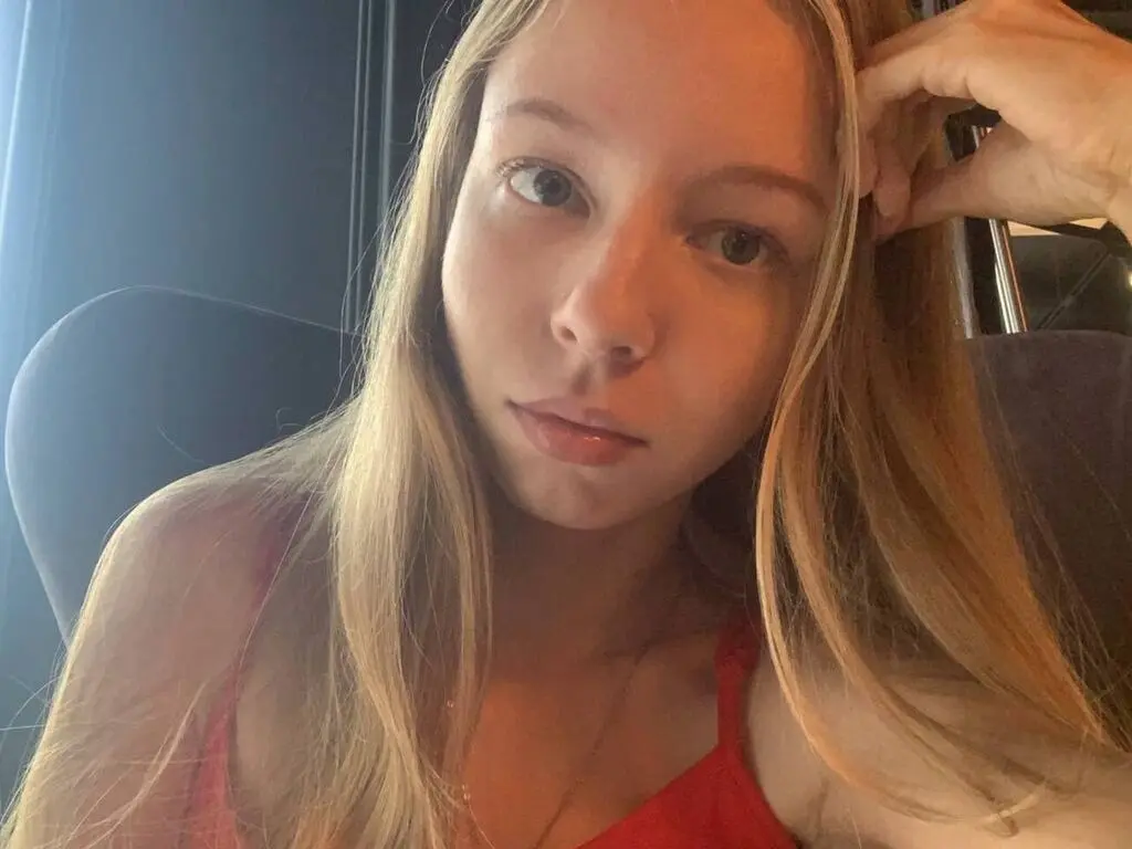 AdenaMcfalls poses with her long blonde hair tucked behind her ear, wearing a red tank top top while looking directly at the camera.