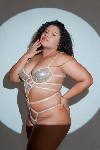 Curvy woman in sparkly silver strappy lingerie posing confidently against a minimalist white circular backdrop.