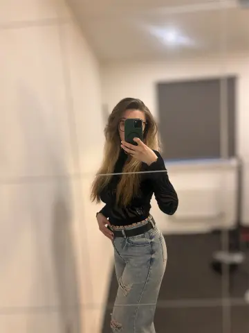Young woman in a black long-sleeve top and light wash denim jeans taking a mirror selfie in a modern room.