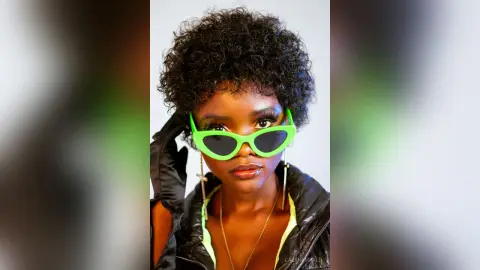 Stylish Black woman with curly hair wearing green sunglasses and a black jacket in a bright studio setting.