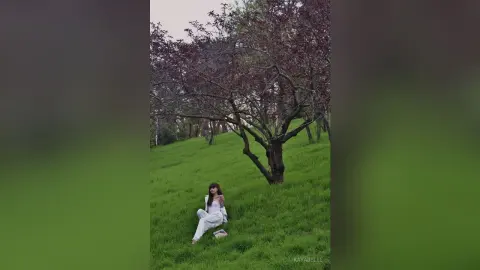 Young woman in a white outfit sitting peacefully on a lush green grass field under a blooming tree.