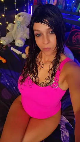 Young woman in a bright pink bodysuit sitting on a bed with a teddy bear in cozy, colorful mood lighting.