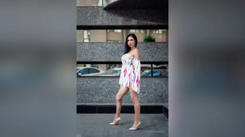 Stylish woman in a white floral mini dress and nude heels posing in front of a modern urban building.