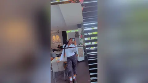 Young woman in white long-sleeve top and patterned trousers taking a mirror selfie in a modern bright room.