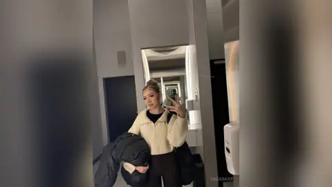 SaraBaier takes a mirror selfie in a white fuzzy sweater and black pants, holding a dark jacket.