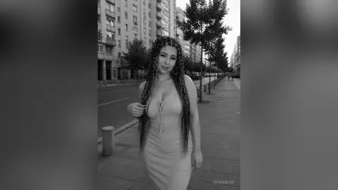 Smiling woman with long braids in a tight dress walking on a city sidewalk in a black and white urban setting.
