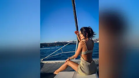 Woman in a beige knit dress relaxing on a sailboat deck against a blue ocean and sunny sky.