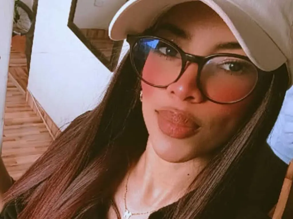 Smiling woman with long brown hair wearing a white baseball cap and black glasses in a bright indoor setting.