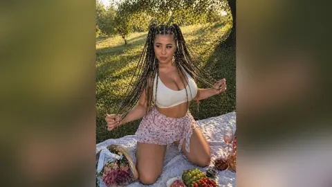 Woman with long braids in a white crop top and floral shorts enjoying a summer picnic in a sunny meadow.