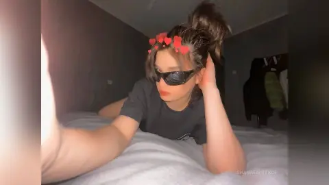 Young woman in a black t-shirt wearing sunglasses while relaxing on a white bed in a cozy bedroom setting.