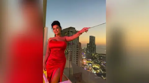 MissyJolie stands on a balcony wearing a red strapless dress against a sunset city skyline.