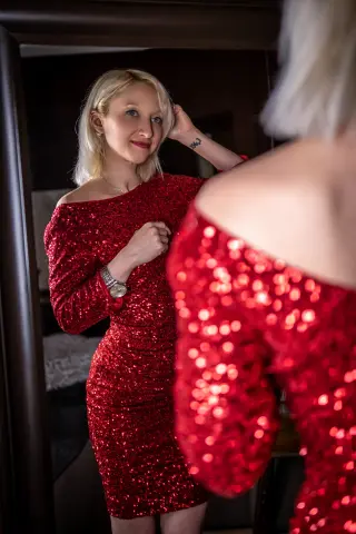 Blonde woman in a sparkling red sequin dress looking at her reflection in a mirror with a soft, elegant mood.