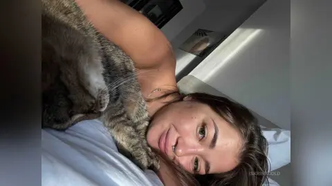Smiling young woman relaxing in bed with a tabby cat in a bright, sunlit bedroom.