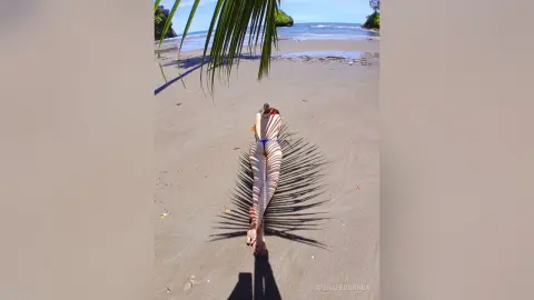 Slim woman in a white striped swimsuit lying on a sandy beach under palm tree shadows on a sunny day.