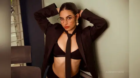 Stylish woman in a black blazer, tie, and bra top posing confidently against a minimalist wall.