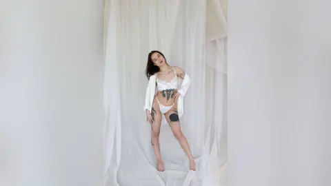 Tattooed woman in white lace lingerie and an open robe posing in a bright, minimalist white studio setting.