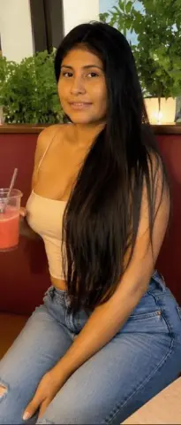 Kendra_Foox poses with long black hair, wearing a tan tank top and blue jeans while sitting in a room with green plants behind her.