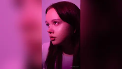 Young woman with dark hair in moody pink lighting looking away thoughtfully.