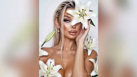 Glamorous blonde woman with glowing skin posing amidst white lilies in a dreamy, ethereal setting.