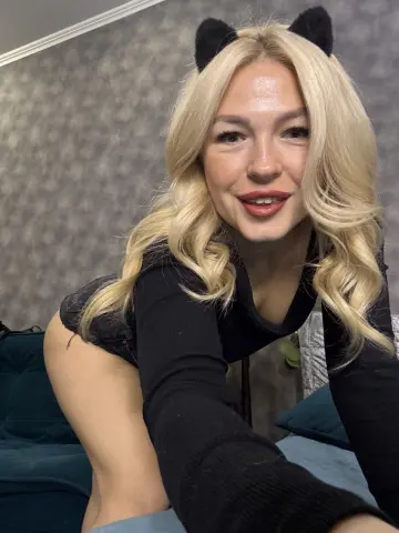 Smiling blonde woman in a black long-sleeve top and cat ears relaxing on a teal sofa in a cozy room.