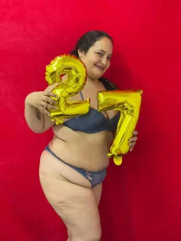 Smiling woman in blue lingerie holding gold number 27 balloons against a red background for a birthday celebration.
